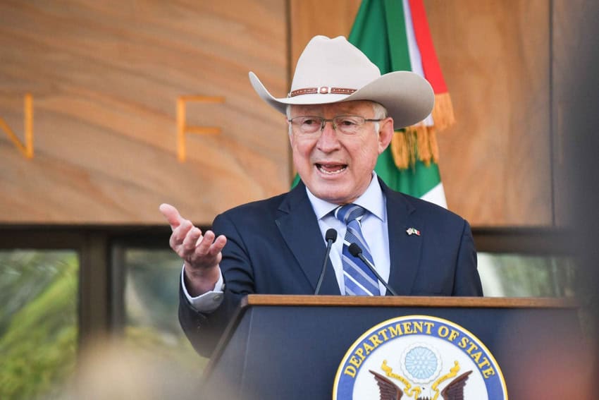 US Ambassasor to Mexico Ken Salazar standing at a US Department of State podium giving a speech and gesturing outward with his right hand. He's wearing a navy blue men's dress suit with a blue, striped tie, and a white cowboy hat.