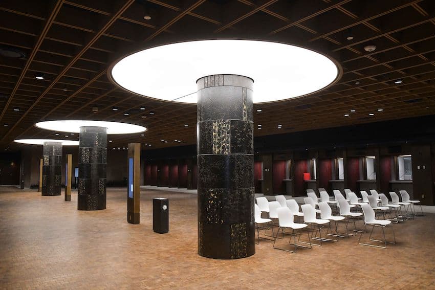 Expansive underground room in the new US Embassy building being finished in Mexico City. There are three rows of temporary metal and plastic chairs set up in the foreground. lluminated service windows run along the far side of the room, while three massive umbrella-like lamps in the photo's foreground, supported by black posts made of Mexican cantera stone, provide the main lighting.