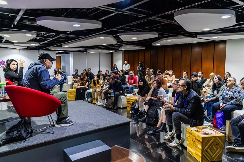 A crowd of young people listen to a poet who sits on a stage reading from his book.